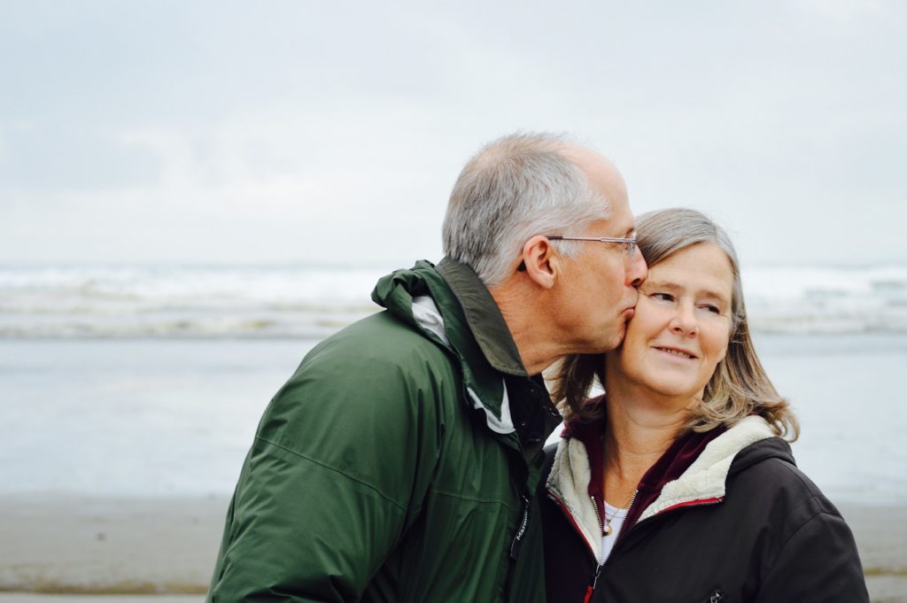 A senior couple standing on the beach. The man is kissing the woman on the cheek, while the woman looks to the left