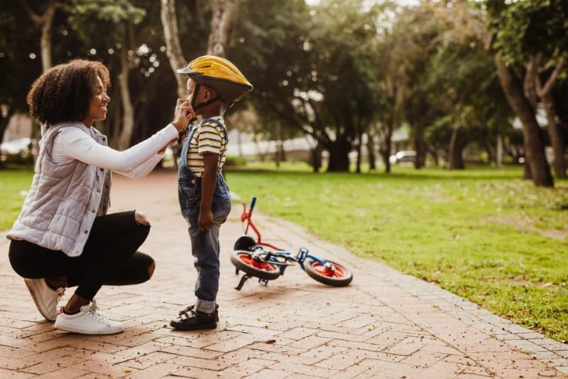 mother helps her son with his helmet while teaching him how to ride a bike
